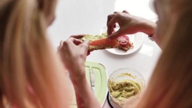 Blonde elinde tost tutar, avokado ezmesi ile yayılmış, ikinci kız üstüne kırmızı balık dilimleri koyar, domates dilimleri ile bir sandviç beyaz bir tabak yatıyor, arkadan görünümü, insanların giysileri pembe.
