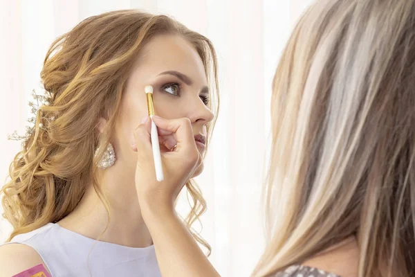 Makeup artist puts makeup on girl model. Brush applies shadows, concealer. beautiful girl model, portrait. Nude colors in makeup. wedding makeup, evening makeup.