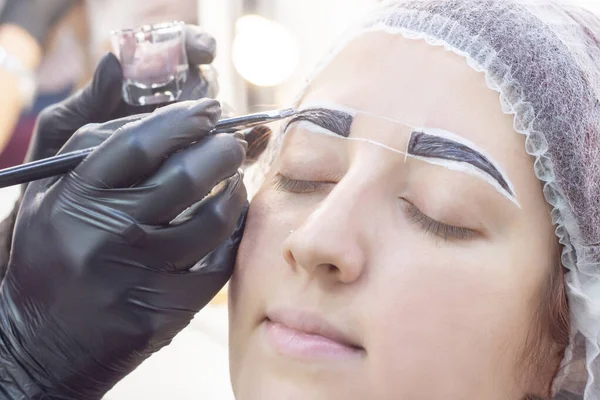 eyebrow dyeing. beauty saloon. the girl lies with her eyes closed on the eyebrow dyeing procedure. The eyebrow master applies brush to the eyebrows of the client.