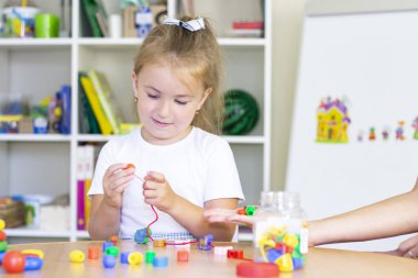 Bir kız çocuğuyla geliştirme ve konuşma terapisi dersleri. Konuşma terapisi egzersizleri ve boncuklarla oyunlar. Kızın ellerinde boncuklar var.
