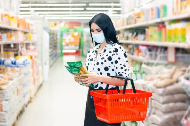 Koruyucu maskeli kız süpermarketten alışveriş yapıyor. Elinde market sepeti ve bakkaliye malzemesi tutan bir kız.