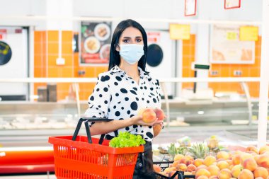 Koruyucu maskeli kız süpermarketten alışveriş yapıyor. Elinde market sepeti olan bir kız meyve seçiyor.