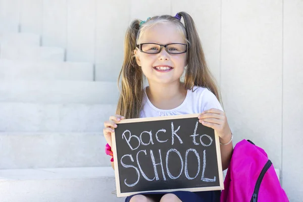 schoolgirl with glasses sits on the stairs, school backpack. Holds a sign in his hands with the inscription back to school