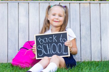 Liseli kız çimenlerin üzerinde oturuyor, okul çantasıyla. Elinde bir tabela tutuyor ve okula geri dönüyor.