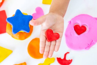 Beyaz bir masanın üzerinde renkli plastikçiden bebek heykelleri. Küçük bir çocuğun eli renkli plastiğin parçalarını sıkıyor. Çocukların yaratıcılığı, eğitici oyunlar, iyi motor beceriler. Plastik kalp.
