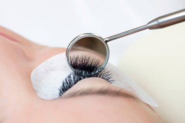 Kirpik uzatma prosedürü. Uzun kirpikli bir kadın gözü. Kapatın. Efendinin ellerinde ayna.