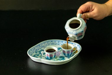 Bir insan çaydanlıktan iki fincana kahve dolduruyor. Bardaklar bir tepside ve kişi çaydanlığı tutuyor.