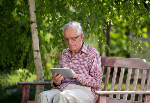 Old man sitting on bench in park and looking at tablet
