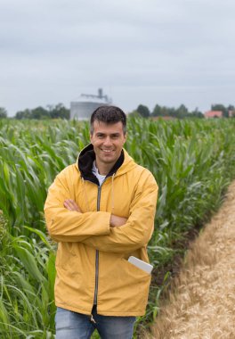 Çapraz kolları ön planda siloları ve Mısır alan ayakta olan yakışıklı bir çiftçi gülümseyen