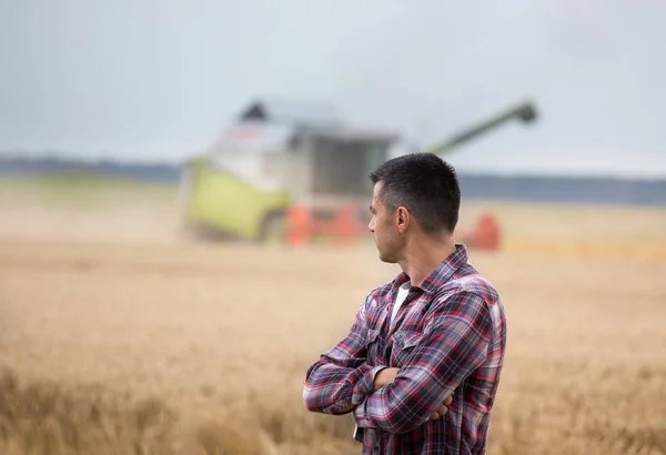 Handsome farmer with crossed arms standing in front of combine harvester during harvest in field
