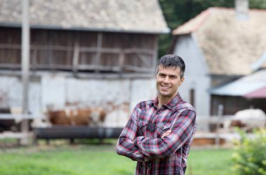 Çapraz kolları simmental inek ve çiftlikte kulübeler önünde ayakta olan kendine güvenen yakışıklı çiftçi