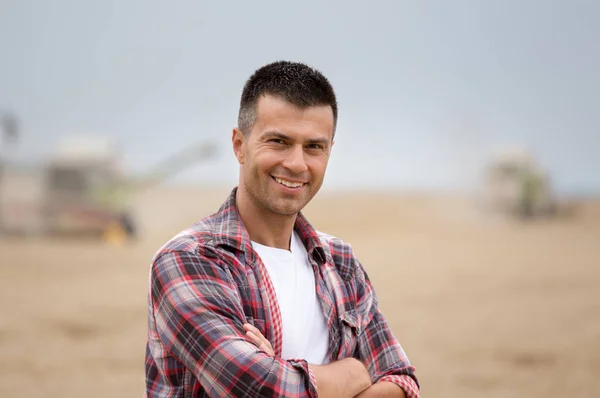 Handsome farmer with crossed arms standing in front of combine harvester during harvest in wheat field