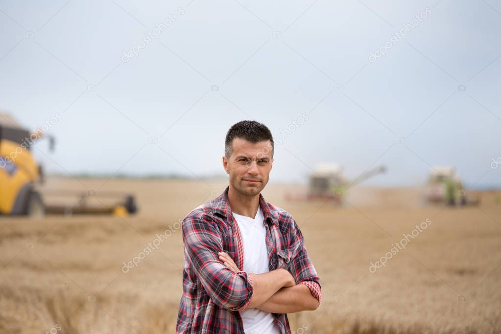 Handsome farmer with crossed arms standing in front of combine harvesters during harvest in wheat field