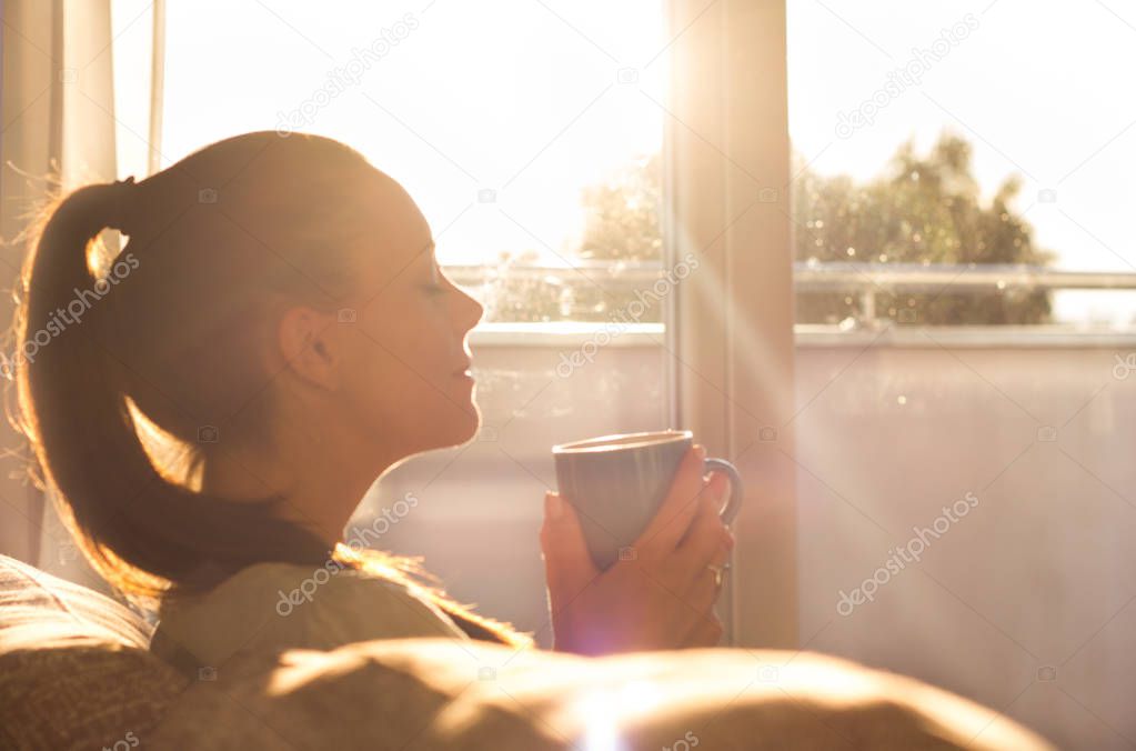 Pretty young woman sitting on sofa and enjoying first morning coffee on sunshine