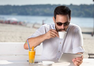 Kafede nehir sahilde oturup, kahve içme ve tablet vasıl seyir güneş gözlüğü ile yakışıklı genç