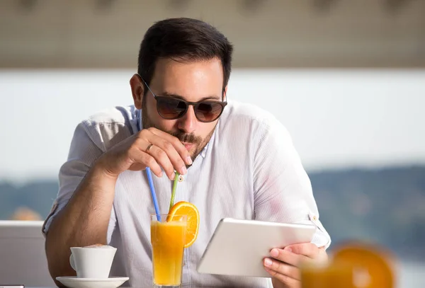 オレンジ ジュースを飲むと ビーチのカフェでタブレットを見てサングラスでハンサムな男は 川の横にある朝を楽しんでください — ストック写真