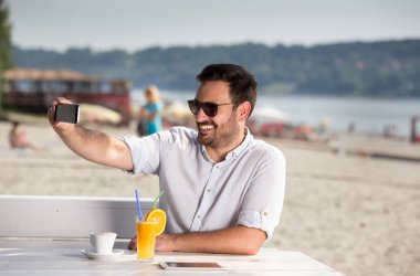 Yakışıklı adam kumsalda web sohbet için telefon kullanarak güneş gözlüklü. Nehir kenarında zevk