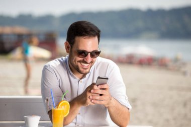 Kahve ve meyve suyu içme ve cep telefonu Beach Cafe bakarak gözlüklü yakışıklı adam. Zevk sabah Nehri yanında