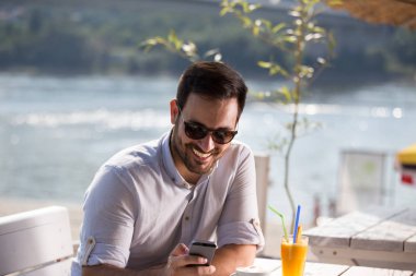 Kahve ve meyve suyu içme ve cep telefonu Beach Cafe bakarak gözlüklü yakışıklı adam. Zevk sabah Nehri yanında