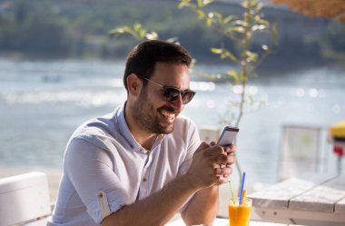 Kahve ve meyve suyu içme ve cep telefonu Beach Cafe bakarak gözlüklü yakışıklı adam. Zevk sabah Nehri yanında