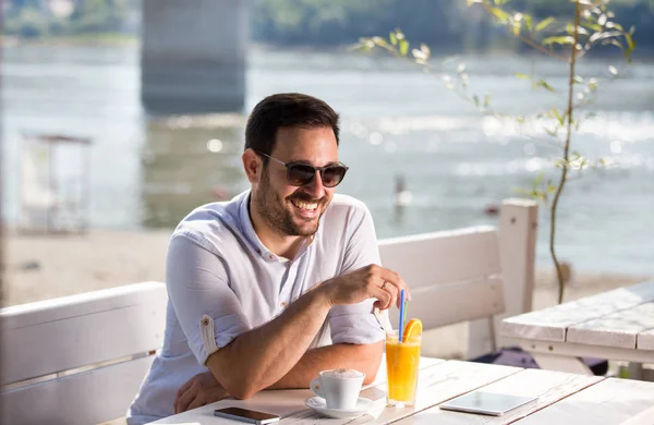 ビーチのカフェでコーヒー ジュースを飲んでのサングラスをかけたハンサムな男は 川の横にある朝を楽しんでください — ストック写真