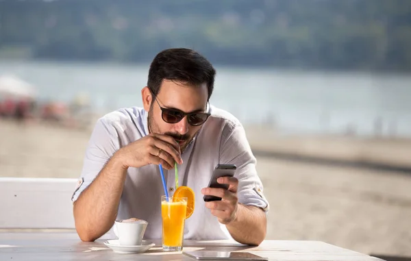 オレンジ ジュースを飲むと ビーチのカフェで携帯電話を見てのサングラスをかけたハンサムな男は 川の横にある朝を楽しんでください — ストック写真