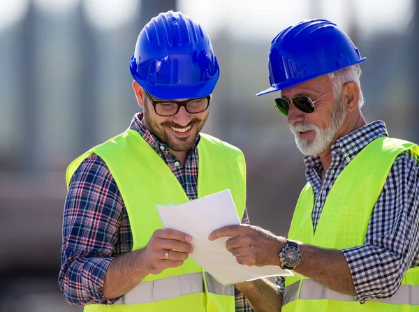 Zwei Zufriedene Ingenieure Betrachten Baupläne Auf Baustelle Mit Metallkonstruktion Hintergrund — Stockfoto