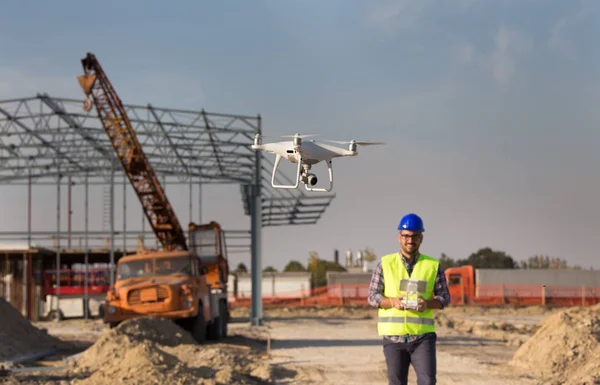 Happy engineer controlling drone above building site with metal construction and crane in background