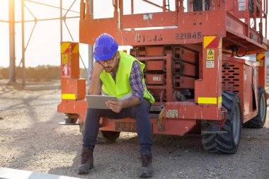 Genç Mühendis vinç üzerinde oturan kask ile tablet ve site oluşturma, düşünme, seyir