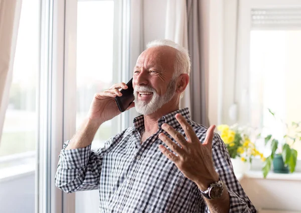 家の窓の横に電話で話して白い髪と髭のシャツを着た興奮した老人 — ストック写真