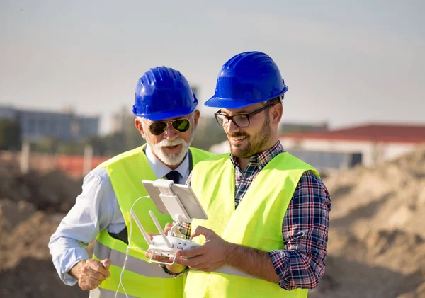 Two engineers with helmets and vests operating with drone by remote control. Technology innovations in construction industry