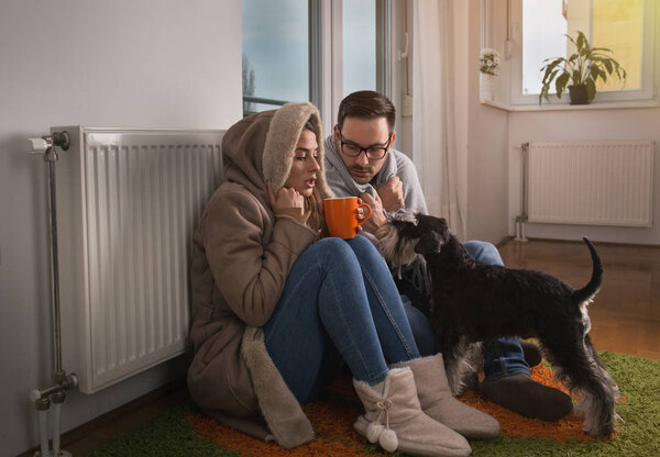 Молодая пара в куртке и покрытая одеялом сидит на полу рядом с радиатором с собакой и пытается согреться
