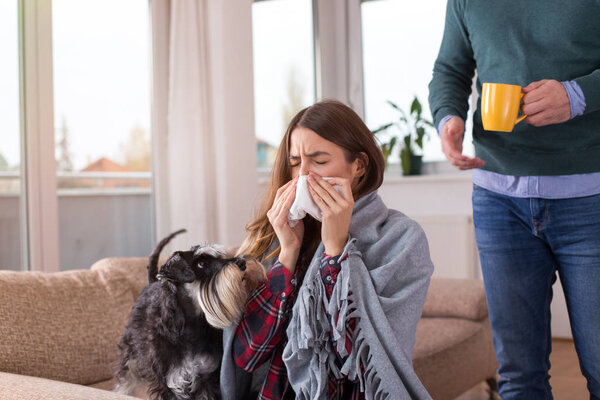Boyfriend bringing tea and taking care of girl having flu. Sitting on sofa at home covered with blanket and blowing nose