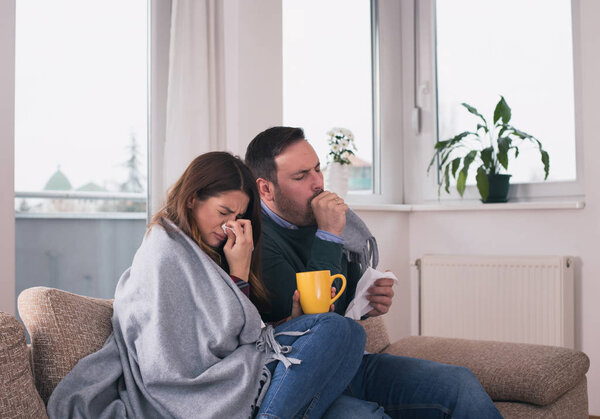 Young couple sitting on sofa covered with blanket, coughing and blowing nose. Cold and flu season 
