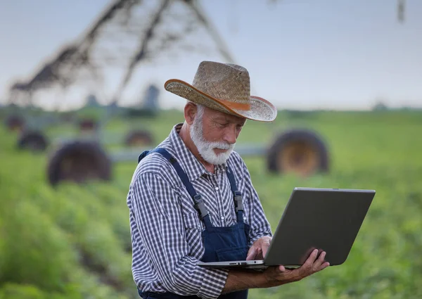 Agricoltore con computer portatile di fronte al sistema di irrigazione in campo — Foto Stock