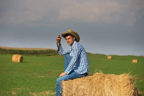 Handsome farmer in plain shirt with straw hat sitting on straw bale roll in field