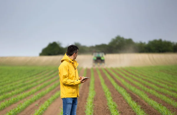 Handsome farmer with tablet standing in corn field in front of tractor in springtime