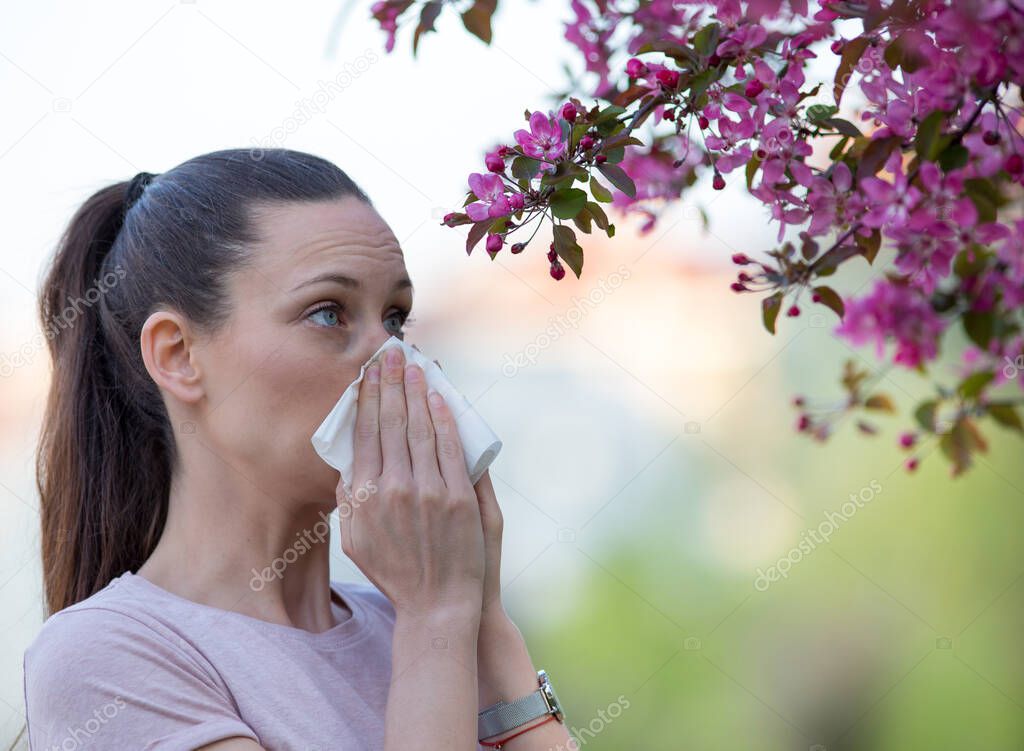 Young pretty woman blowing nose in front of blooming tree. Spring allergy concept