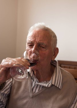 Gömlek ve yelek giyen kıdemli adam evde camdan kırmızı şarap içiyor.