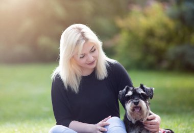 Baharda çimenlerde köpeğiyle oturan sarışın genç kızın portresi. Minyatür Schnauzer köpeğiyle.