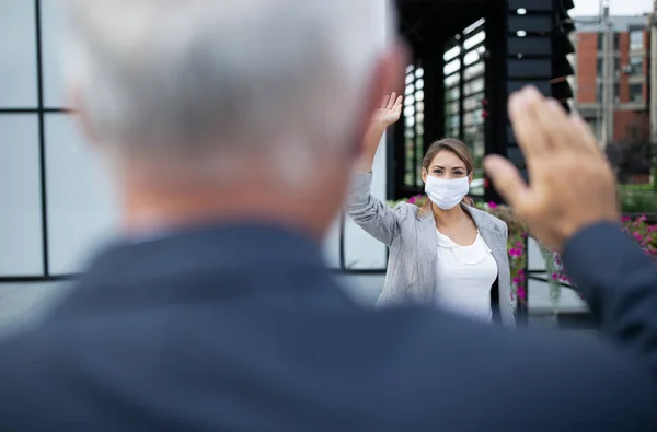 Business woman with safety mask and man greeting by waving hands in front of office building. Virus protection and social distancing concept at work