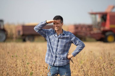 Soya tarlasında durup güneşe bakarken arka planda çalışan karavanla hasat ve traktörü birleştiren yakışıklı çiftçi.
