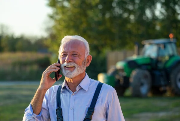 Portret Zadowolonego Starszego Rolnika Rozmawiającego Przez Telefon Komórkowy Przed Ciągnikiem — Zdjęcie stockowe