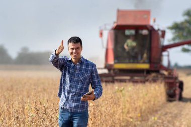 Soya tarlasında duran ve arka planda çalışan hasatçıyı birleştirirken baş parmağını gösteren tabletli yakışıklı bir çiftçi.
