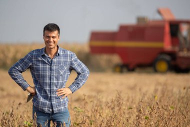 Soya tarlasında duran ve elinde tablet tutan yakışıklı çiftçi arka planda çalışan hasatçıyı birleştiriyor.