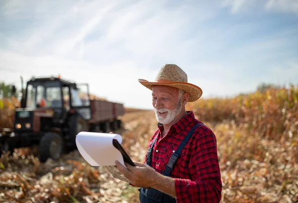 年长的农民头戴草帽 手里拿着笔记本 站在拖拉机前 拖着装满玉米芯的拖车收割庄稼 — 图库照片