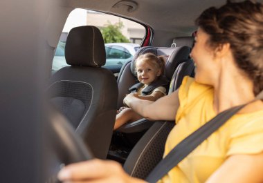 Anne, güneşli bir günde arka koltukta oturan ve göz teması kuran küçük çocuğu, sürücü koltuğunda otururken geriye bakıyor.