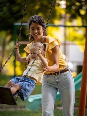Çocuk parkında sallanıyor. Neşeli genç anne kızının salıncakta durup eğlenmesine yardım ediyor. 