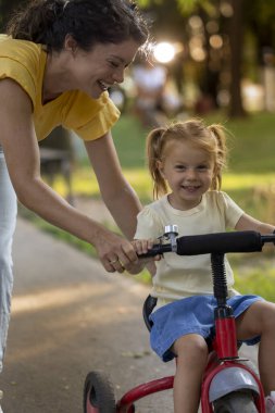 Mutlu annesinin yardımıyla parkta çarklı bisiklete binen sevimli, gülümseyen bir çocuk.