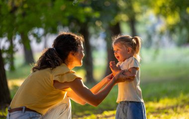 Anne, güneşli bir parkta çocuğuyla neşeyle meşgul oluyor. Huzurlu doğal ortamda kahkaha ve iletişim anı yaratıyor.